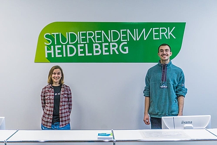 Links steht Carola Kasimir vor dem grünen Logo des Studierendenwerks Heideöberg. Neben ihr aud der rechten Seite lächelt Raphael Wankelmuth in die Kamera.