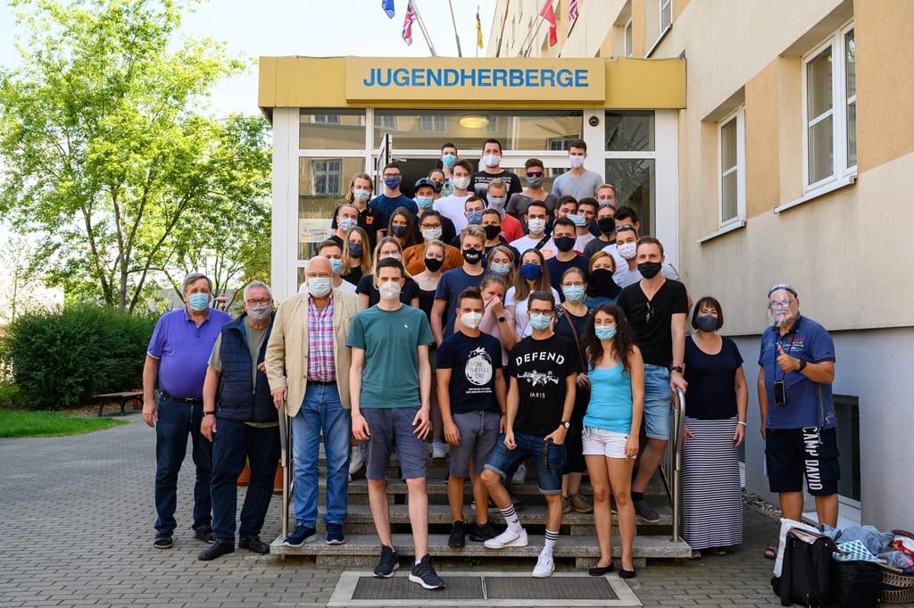 Gruppenfotos von Studierende auf einer Treppe vor einer Jugendherberge. Alle abgebildeten Studierenden tragen einen Mund-Nasen-Schutz.